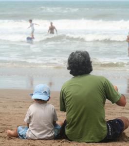 A Father sitting on the beach with his small child, watching the ocean surf.